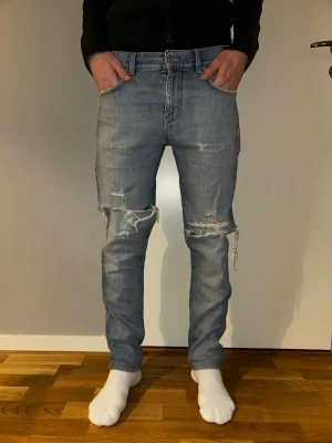 Slim J.Lindberg jeans - Säljer ett par ljusblå slim jeans med slitningar och hål vid knäna. Jeansen har klassisk femficksmodell, normal midja och är tillverkade i bomull med lite stretch. Perfekta för en avslappnad och trendig look. 32/32