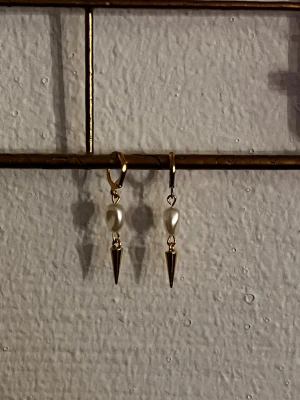Guldörhängen med pärla och spike - Snygga örhängen i guldton med oval vit pärla och hängande guldfärgad spike. Trendig design som kombinerar klassisk pärla med edgy detaljer. Perfekta för dig som gillar att sticka ut med dina accessoarer.