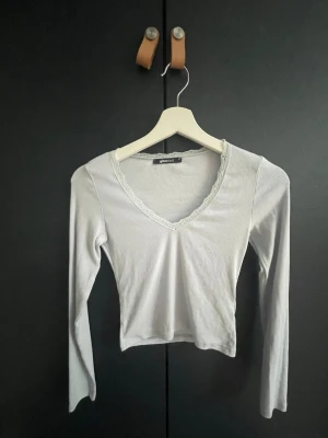 Ljusgrå croppad topp med spets - Supersöt ljusgrå med lång ärm och v-ringning. Tunn ribbad bomullsmix och spetsdetalj runt halsen ger en extra fin touch. Perfekt till jeans eller kjol för en trendig look.
