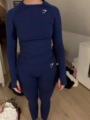 Mörkblått träningsset från Gymshark - Säljer ett mörkblått träningsset från Gymshark med långärmad topp och matchande tights. Setet är tight fit, har vita loggor på både tröja och byxa och är gjort i ett stretchigt funktionsmaterial som passar perfekt till gymmet eller löpning.