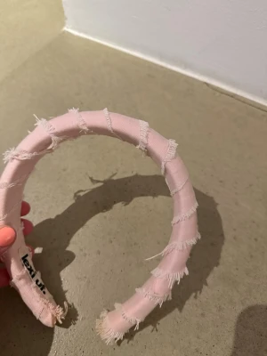 Ljusrosa diadem med fransiga kanter - Säljer ett ljusrosa diadem från Lexi uf med trendiga fransiga kanter. Diademet är vadderat och har en bred form som ger en cool och modern look. Perfekt accessoar för att lyfta din outfit med en söt och edgy touch.