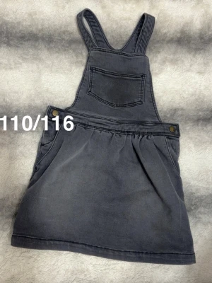 Mörkgrå jeanskjol med hängslen - Snygg mörkgrå jeanskjol med hängslen och bröstficka. Kjolen har två knappar i sidorna och en klassisk jeanslook. Perfekt för en avslappnad och trendig stil.🌸 Fint använt skick. Strechigt och mjukt material. Storlek 110/116 