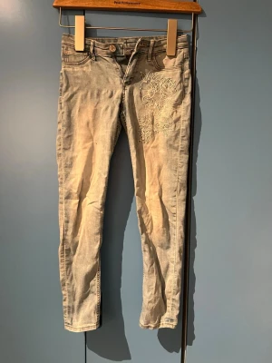 Ljusblå skinny jeans med broderi - Snygga ljusblå skinny jeans med blommigt broderi på ena låret. Jeansen har klassisk femficksdesign och är tillverkade i mjukt denimtyg. Perfekta för dig som gillar detaljer och vill sticka ut lite extra.