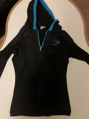 Svart sporthoodie från Gymshark - Snygg svart sporthoodie från Gymshark med blå detaljer och dragkedja. Tröjan har lång ärm, huva med blå kant och ett geometriskt mönster framtill. Perfekt för träning eller chill. Materialet är stretchigt och andas bra.