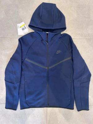 Blå Nike hoodie med dragkedja - Snygg blå hoodie från Nike med hel dragkedja och huva. Hoodien har en stilren design med diskret Nike-logga på bröstet och svarta detaljer längs dragkedjan och ärmsluten. Tillverkad i mjukt och stretchigt material som känns bekvämt. Aldrig använd!