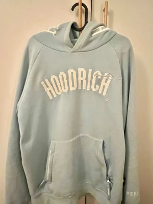 Ljusblå hoodie från Hoodrich - Snygg ljusblå hoodie från Hoodrich med stor vit logga framtill och tryck på ryggen. Hoodien har huva med broderade detaljer och en stor magficka med dragkedja. 