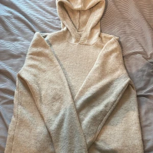 Beige hoodie i stickat material - Mysig beige hoodie i stickat material med huva och långa ärmar. Tröjan har en enkel och clean look utan tryck eller mönster, perfekt för dig som gillar en avslappnad stil. Passformen är normal och den är riktigt skön att ha på sig. Bra för kallare dagar. 