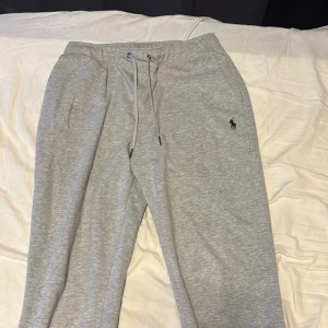 Grå mjukisbyxor från Polo Ralph Lauren - Snygga grå mjukisbyxor från Polo Ralph Lauren med dragsko i midjan och broderad logga på vänster ben. Byxorna har ribbade muddar vid bensluten och är tillverkade i mjuk bomullsmix för maximal komfort. Perfekta för chill dagar eller när du vill vara extra bekväm.