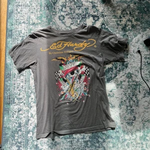 Grå Ed Hardy t-shirt med tryck - Snygg grå t-shirt från Ed Hardy med stort färgglatt tryck framtill, prydd med paljetter och coolt dödskallemotiv. Klassisk rund halsringning och korta ärmar. Logga i gult både fram och bak. Perfekt för dig som gillar statement-plagg.