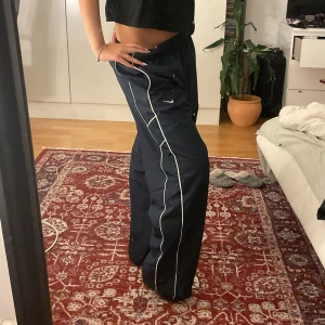 Marinblå Nike täckbyxor med vita detaljer - Säljer ett par marinblå täckbyxor från Nike med vita kontrastsömmar längs benen. Byxorna har en loose passform, elastisk midja och är perfekta för dig som gillar sportig stil. Materialet är syntet och de har en klassisk streetkänsla. Strl S/M för mig för dem ska vara baggy.