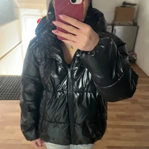 Svart pufferjacka - Säljer en svart, glansig pufferjacka från Vila i storlek 36 som är slutsåld. Jackan har en oversized look, dragkedja framtill och stor huva. Perfekt för kalla dagar och ger en cool vibe till din outfit.