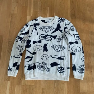 Vit stickad tröja med svarta motiv Mouli - Vit stickad tröja från Mouli med svarta grafiska motiv som diamanter, cyklar, helikoptrar och smileys. Tröjan har rund halsringning, ribbade muddar och lång ärm. Perfekt för dig som gillar unika och lekfulla plagg.