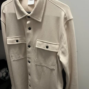 Beige overshirt från Selected Homme - Snygg beige overshirt från Selected Homme med två stora bröstfickor och svarta knappar framtill. Overshirten har en klassisk krage och är tillverkad i ett kraftigare bomullstyg. Perfekt att slänga över en t-shirt för en clean look.