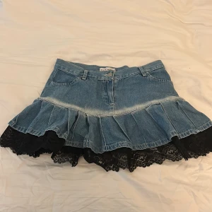 Jeanskjol med volang och spets No Gimmick - Supersöt jeanskjol från No Gimmick med volangkant och svart spets nertill. Kjolen är kort, har klassiska jeansfickor fram och bak samt bälteshällor. Perfekt för dig som gillar en mix av denim och romantiska detaljer.