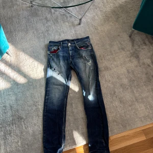 Replay blå jeans med slitningar - Säljer ett par blå jeans från Replay med coola slitningar och lagningar framtill. Jeansen har raka ben, klassiska bakfickor och detaljer i rött vid fickorna. Materialet är jeans i bomull och de har en snygg tvättad look.