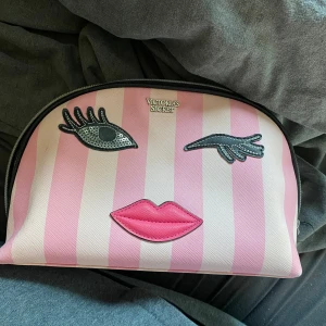 Victoria's Secret Sminkväska - Superfin necessär från Victoria's Secret med rosa och ljusrosa ränder. Framsidan har ett coolt ansikte med applikationer i form av ögonfransar, öga och läppar. Väskan har dragkedja och är perfekt för smink eller småprylar.