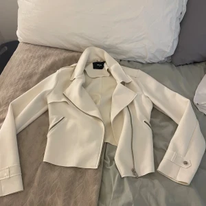 Vit croppad kavajjacka från Bik Bok - Snygg vit croppad kavajjacka från Bik Bok med dragkedjor i silver på sidorna och klassisk krage. Jackan har långa ärmar och stilrena detaljer vid ärmsluten. Perfekt för dig som vill ha en clean och modern look.