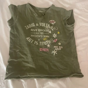Grön t-shirt Zadig & Voltaire tryck - Olivgrön t-shirt från Zadig & Voltaire med coolt tryck framtill med blommor, stjärnor, peace-märken och text. Rund hals med råa kanter och knappdetalj. Skön passform och tillverkad i mjuk bomull. Perfekt för en avslappnad och trendig look. Ett litet hål på bröstet (kolla sista bilden för närbild)storlek 134-140 men passar mig som är 159 cm perfekt 💖💓💕
