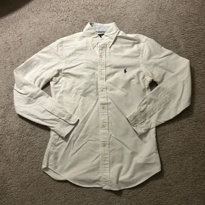 Vit skjorta från Ralph Lauren slim fit - Vit långärmad skjorta från Ralph Lauren i slim fit-modell. Klassisk button-down krage, broderad marinblå logga på bröstet och knappar framtill. Tillverkad i mjuk bomull, perfekt för en clean och stilren look.