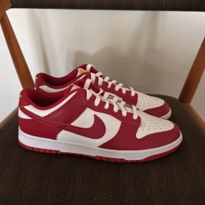 Nike Dunk Low Retro Gym Red/White - Nike Dunk Low Retro i färgen Gym Red/White. Klassiska sneakers med vit bas och röda overlays, gul broderad Nike-logga på hälen och vit sula med röd yttersula. Skorna har snörning och är tillverkade i skinn för en clean och tidlös look. Skorna är helt nya och i storlek 44. Hör av er vid frågor och funderingar. 