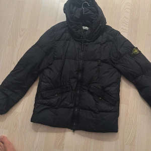 Svart pufferjacka från Stone Island - Säljer en svart pufferjacka från Stone Island med huva och klassisk patch på ärmen. Jackan har två stora fickor framtill med lock och dragkedja. Perfekt för kalla dagar och har en clean, stilren look.