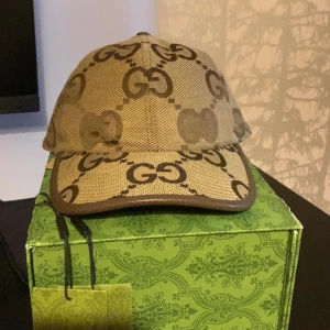 Beige keps från Gucci med GG-mönster - Snygg beige keps från Gucci med det klassiska GG-logomönstret i brunt över hela kepsen. Kepsen är tillverkad i bomullscanvas och har bruna detaljer längs skärmen och kanterna. Perfekt accessoar för att lyfta din outfit med lyxig streetstyle.