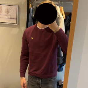 Vinröd sweatshirt från Lyle & Scott - Säljer en vinröd sweatshirt från Lyle & Scott med klassisk rund hals och gul logotyp på bröstet. Tröjan har långa ärmar, ribbade muddar och är gjord i mjuk bomull. Perfekt för chill dagar och enkel att matcha med jeans eller joggers.