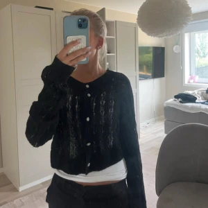 Svart kofta från Lindex med pärlknappar - Säljer en svart, stickad kofta från Lindex med fina pärlknappar. Koftan är croppad, har lång ärm och ett genombrutet mönster som ger en luftig känsla. Perfekt att slänga över en topp för en snygg layering-effekt.