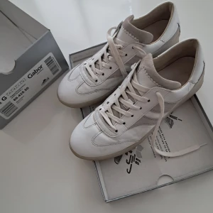 Vita sneakers från Gabor i skinn - Säljer ett par fräscha vita sneakers från Gabor i storlek 40. Skorna är gjorda i nappaskinn och mocka, med snygga detaljer och klassisk snörning. Sulan är i gummi och har en låg kilklack på 15 mm. Perfekta för dig som gillar stilrena och bekväma sneakers.
