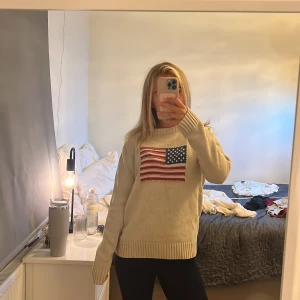 Beige stickad tröja från GANT med flagga - Säljer en beige stickad tröja från GANT i storlek small, tillverkad i 100% bomull. Tröjan har ett broderat amerikanskt flaggmotiv på bröstet och ribbade muddar vid ärmslut och nederkant. Perfekt för dig som gillar preppy stil och snygga detaljer.