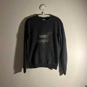 Svart Saint Laurent sweatshirt - Svart sweatshirt från Saint Laurent med coolt tryck på bröstet och blixtdetalj. Tröjan har ribbad rund hals och muddar vid ärmslut och nederkant. Materialet är mjuk bomull och insidan är frottéliknande för extra komfort. Perfekt för dig som gillar streetstyle.