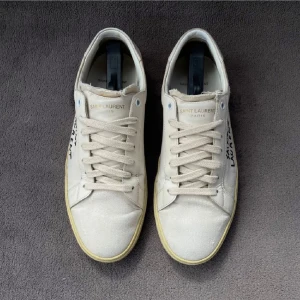 Saint Laurent vita sneakers i canvas - Snygga vita sneakers från Saint Laurent i canvas med handskriven logga på sidan. Skorna har snörning, rund tå och platt sula i beige ton. Perfekta för dig som gillar minimalistisk och stilren design med en twist.