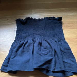 Marinblå bandeau tubtopp med volang - Säljer en marinblå bandeau tubtopp med smockad överdel och volang nertill. Toppen är axelbandslös och har en söt rynkad kant upptill. Perfekt för varma dagar eller att styla lager på lager.