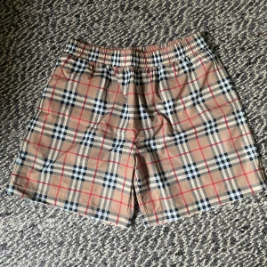 Rutig Burberry shorts  - Snygga shorts från Burberry i klassiskt beige, svart, vit och röd rutig design. De har elastisk midja och är tillverkade i 100% bomull, vilket gör dem både bekväma och lätta. Perfekta för sommardagar när du vill ha en clean och stilren look.