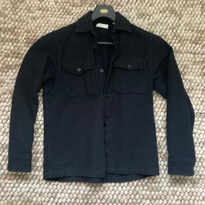Svart overshirt med fickor - Säljer en svart overshirt med klassisk krage och två stora bröstfickor med knappar. Skjortan har långa ärmar och stängs med knappar framtill. Perfekt för lager-på-lager och en clean look.