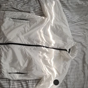 Vit vindjacka från Calvin Klein XL - Snygg vit vindjacka från Calvin Klein i storlek XL. Jackan är tillverkad i glansig polyester, har svarta dragkedjor, två fickor och en hög krage. Vatten- och vindavvisande material, perfekt för blåsiga dagar. Diskret CK-logga på ärmen.
