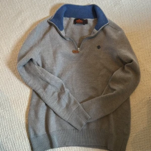 Grå merinoulltröja från Morris - Snygg grå tröja från Morris i 100% merinoull med halv dragkedja och blå insida på kragen. Liten broderad logga på bröstet och ribbade muddar. Perfekt för lager-på-lager och chillare dagar.