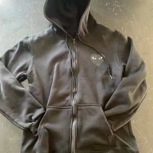 Svart hoodie med hjärtdetalj - Svart hoodie från CDGmed dragkedja, huva och fickor framtill. På bröstet sitter det ikoniska svarta hjärtat med ögon. Perfekt för dig som gillar streetstyle och vill ha något stilrent men ändå unikt.