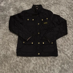 Svart quiltad jacka Barbour XL - Svart quiltad jacka från Barbour International i storlek XL. Jackan har guldiga knappar, fyra fickor framtill och Barbour-logga på bröstet. Klassisk krage och snyggt mönstrat foder. Perfekt för dig som vill ha en stilren och tidlös look.