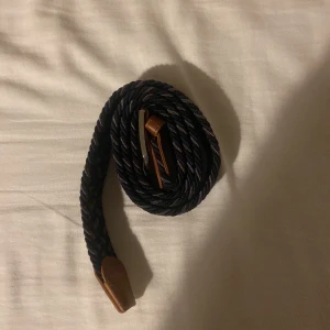 Mörkblått flätat bälte med läderdetaljer - Snyggt mörkblått flätat bälte med bruna läderdetaljer och silverfärgat spänne. Bältet har en stilren och tidlös look med en twistad struktur som ger extra karaktär. Perfekt för att lyfta din outfit med en klassisk accessoar.