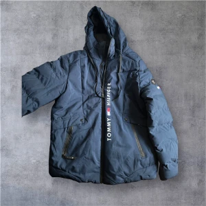 Blå pufferjacka från Tommy Hilfiger - Snygg marinblå pufferjacka från Tommy Hilfiger med huva och dragkedja framtill. Jackan har stora fickor med dragkedja och logotypen tryckt längs dragkedjan. Perfekt för kalla dagar och har en modern, loose passform.