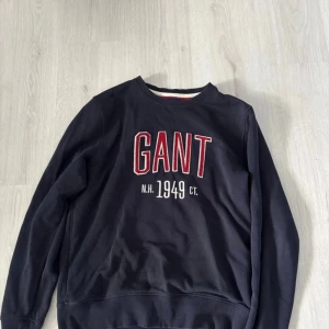 Mörkblå sweatshirt från GANT, storlek L - Mörkblå sweatshirt från GANT med stor röd och vit broderad logga på bröstet. Rund hals och långa ärmar, insida i rött tyg. Tillverkad i mjuk bomull för skön känsla. Perfekt för dig som gillar klassisk college-stil.