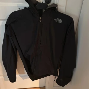 Svart vindjacka från The North Face - Säljer en svart vindjacka från The North Face med huva och dragkedja. Jackan har en stilren design och är perfekt för blåsiga dagar. Den har långa ärmar och är tillverkad i ett lätt material som skyddar mot vind. Drags kedjan är trög på jackan men annars i bra skick