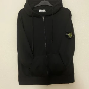 Svart hoodie från Stone Island - Svart hoodie från Stone Island med dragkedja framtill, huva med snörning och klassisk logotyp-patch på ärmen. Tröjan har känguruficka och är tillverkad i mjuk bomull. Perfekt för en avslappnad och stilren look.