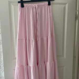 Ljusrosa volangkjol med snörning - Supersöt ljusrosa långkjol med volanger och snörning i midjan. Kjolen har en luftig och flowy känsla, perfekt för varma dagar. Tillverkad i ett lätt, strukturerat tyg som ger en avslappnad men ändå trendig look.