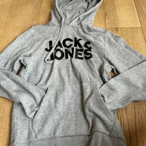 Grå hoodie från Jack & Jones - Grå hoodie från Jack & Jones med svart logga framtill. Tröjan har huva med snörning och en stor magficka. Tillverkad i mjuk bomullsmix, perfekt för chill dagar. Klassisk passform och långa ärmar.