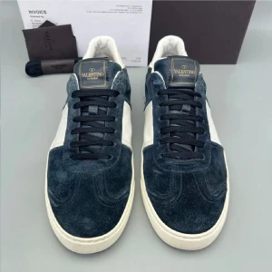 Valentino Garavani sneakers blå/vit - Snygga Valentino Garavani sneakers i mörkblå mocka med vita skinnpaneler och vit sula. Klassisk låg modell med svarta snören och ikoniska nitar på hälen. Perfekta för dig som vill ha en exklusiv och stilren look.