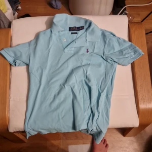 Ljusblå Polo Ralph Lauren t-shirt M - Snygg ljusblå t-shirt från Polo Ralph Lauren i klassisk pikémodell med krage och korta ärmar. Ikoniska lilla loggan broderad på bröstet. Skön passform och tillverkad i bomull, perfekt för en chill och stilren look.