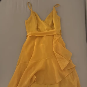 Gul omlottklänning med volanger - Superfin gul kortklänning med tunna axelband och omlottliv. Klänningen har volangdetaljer och ett skönt, lätt tyg som ger en flowig känsla. Perfekt för varma dagar när du vill sticka ut med en färgglad look.