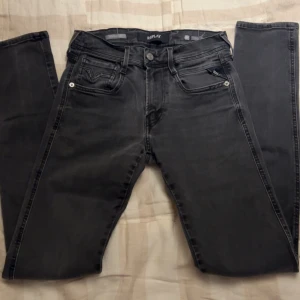 Replay Anbass svarta jeans, stl 27 - Snygga svarta Replay Anbass jeans i klassisk femficksmodell. Jeansen har en mörk tvätt med diskreta slitningar och är slim fit. Märkesdetaljer syns på fickorna och bak med läderpatch. Perfekta för dig som gillar en stilren look.